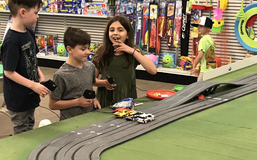 Three children hold controllers with concentration and excitement on their faces as they race colorful slot cars on a track, with a toy-filled background capturing the lively atmosphere of the play area.