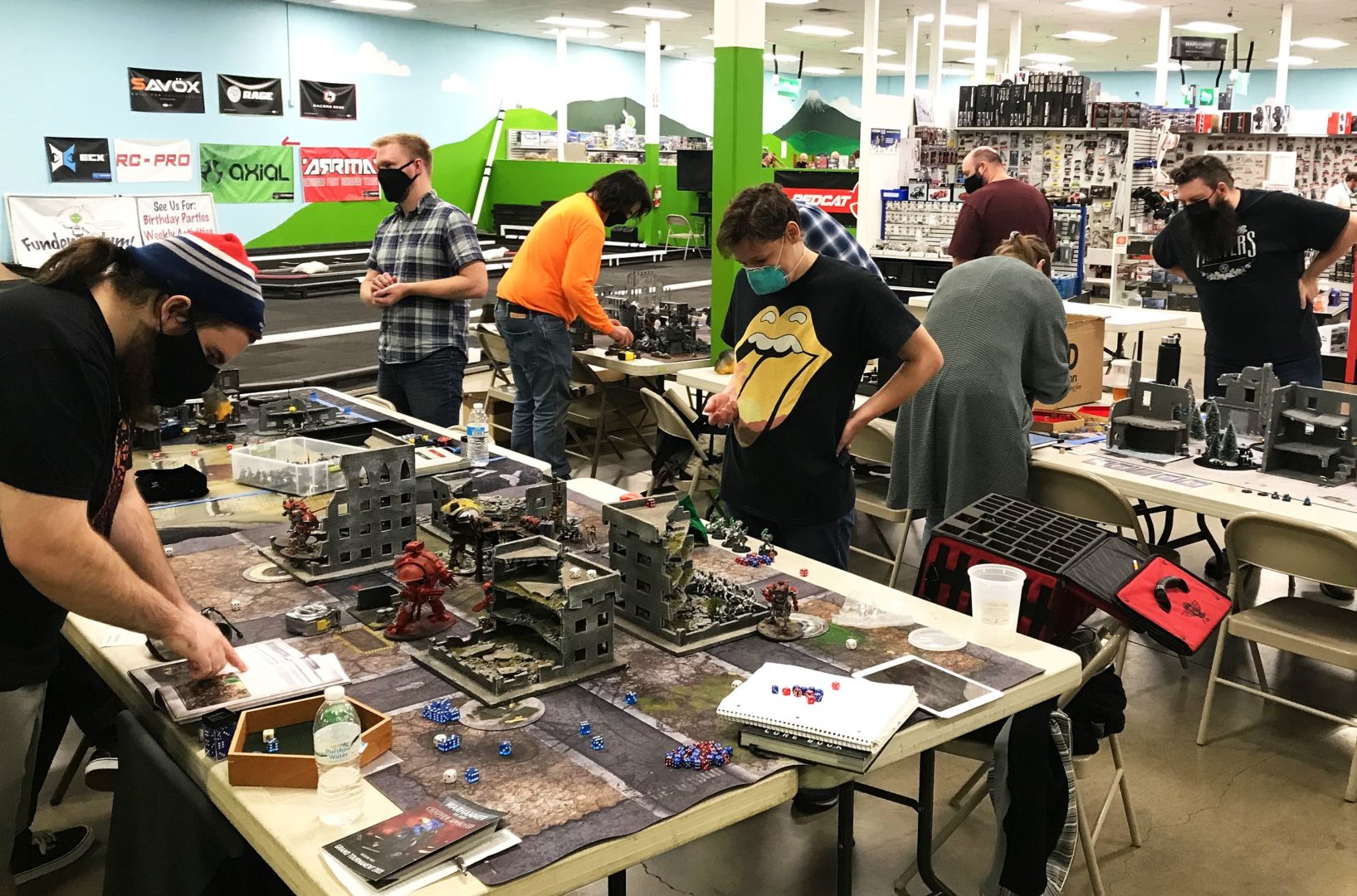 Enthusiasts gather around a table for a Warhammer game, deeply engaged in strategic play amid a detailed miniature battlefield setup, with various painted figures and terrain pieces in a hobby gaming store