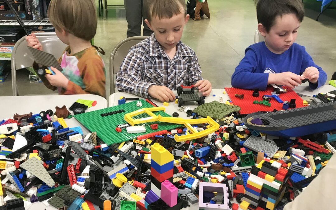 Three young boys are deeply engrossed in building with LEGOs, their small hands selecting from a vibrant and chaotic assortment of blocks on the table, as they create imaginative structures and vehicles