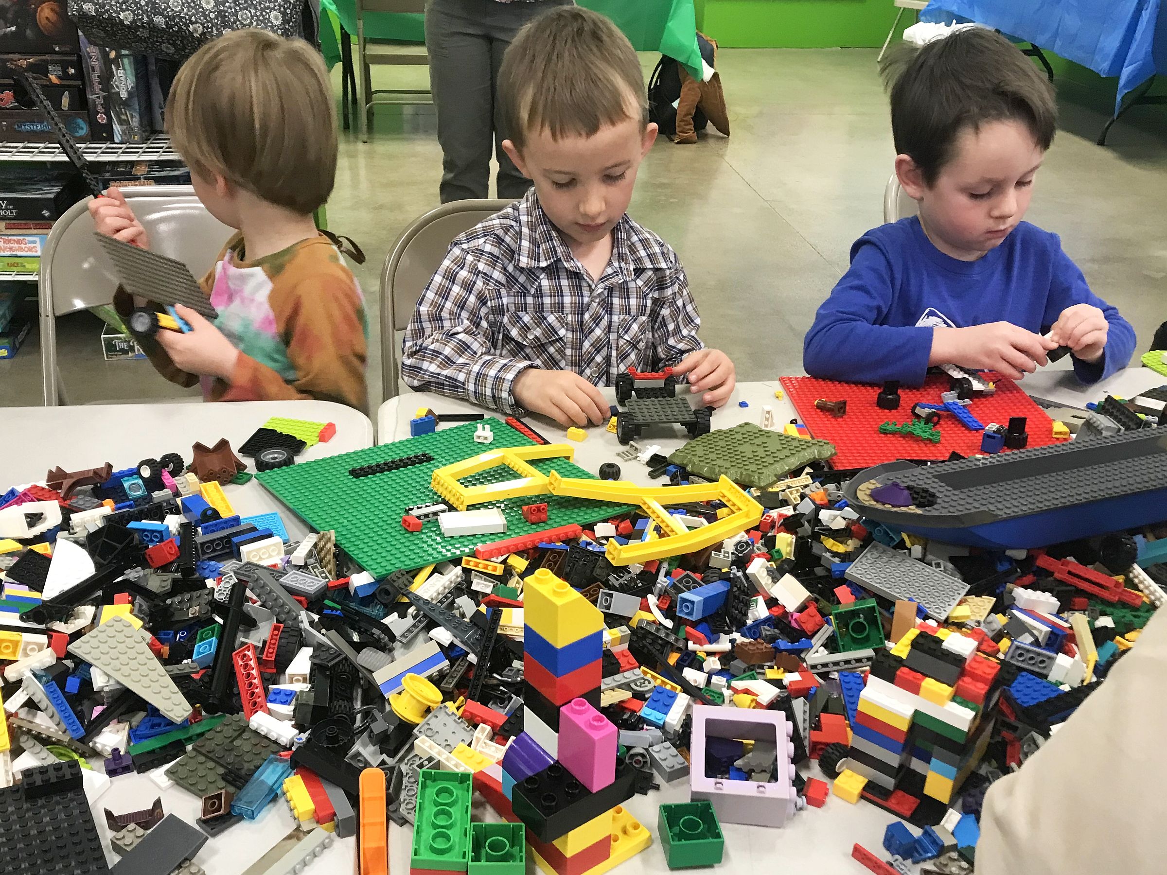 Three young boys are deeply engrossed in building with LEGOs, their small hands selecting from a vibrant and chaotic assortment of blocks on the table, as they create imaginative structures and vehicles