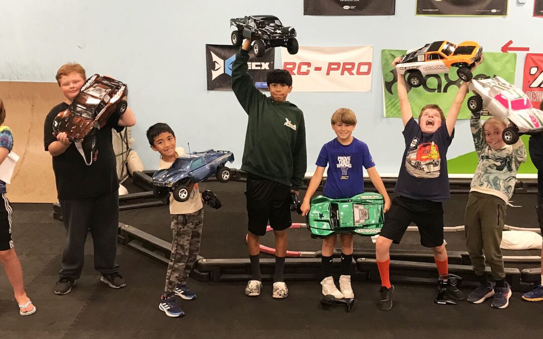 A group of seven jubilant children raise their custom-painted RC cars triumphantly in the air, celebrating their creativity and engineering skills in a play area with RC racing brands displayed in the background.