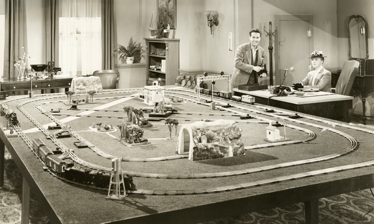an old photograph of a man standing and a woman sitting at a table covered with a model train set