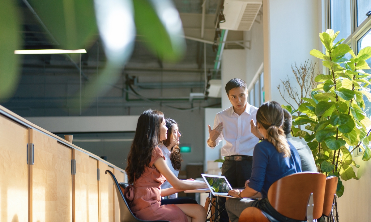 a working team seated at a table together and looking at a standing man who is explaining something